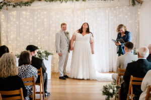 Bride and groom stand hand in hand laughing during the ceremony